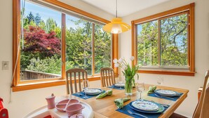 Zona pranzo con tavolo in legno apparecchiato per due, ampie finestre vista alberi e giardino, e una lampada a sospensione gialla.