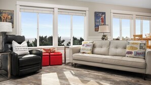 A living room with a beige sofa, a red ottoman, a black leather chair, and a glass coffee table. There are two windows with blinds, a painting on the wall, and a lamp on the side table. - Casa de Marea - 2BD Condo - Close to Beach +Garage (Lincoln City)