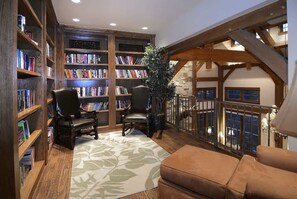 A library with bookshelves, two chairs, a patterned rug, and a balcony with a railing.