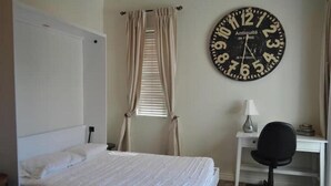 A bedroom with a bed, a desk, a chair, a wall clock, and a window with curtains.
