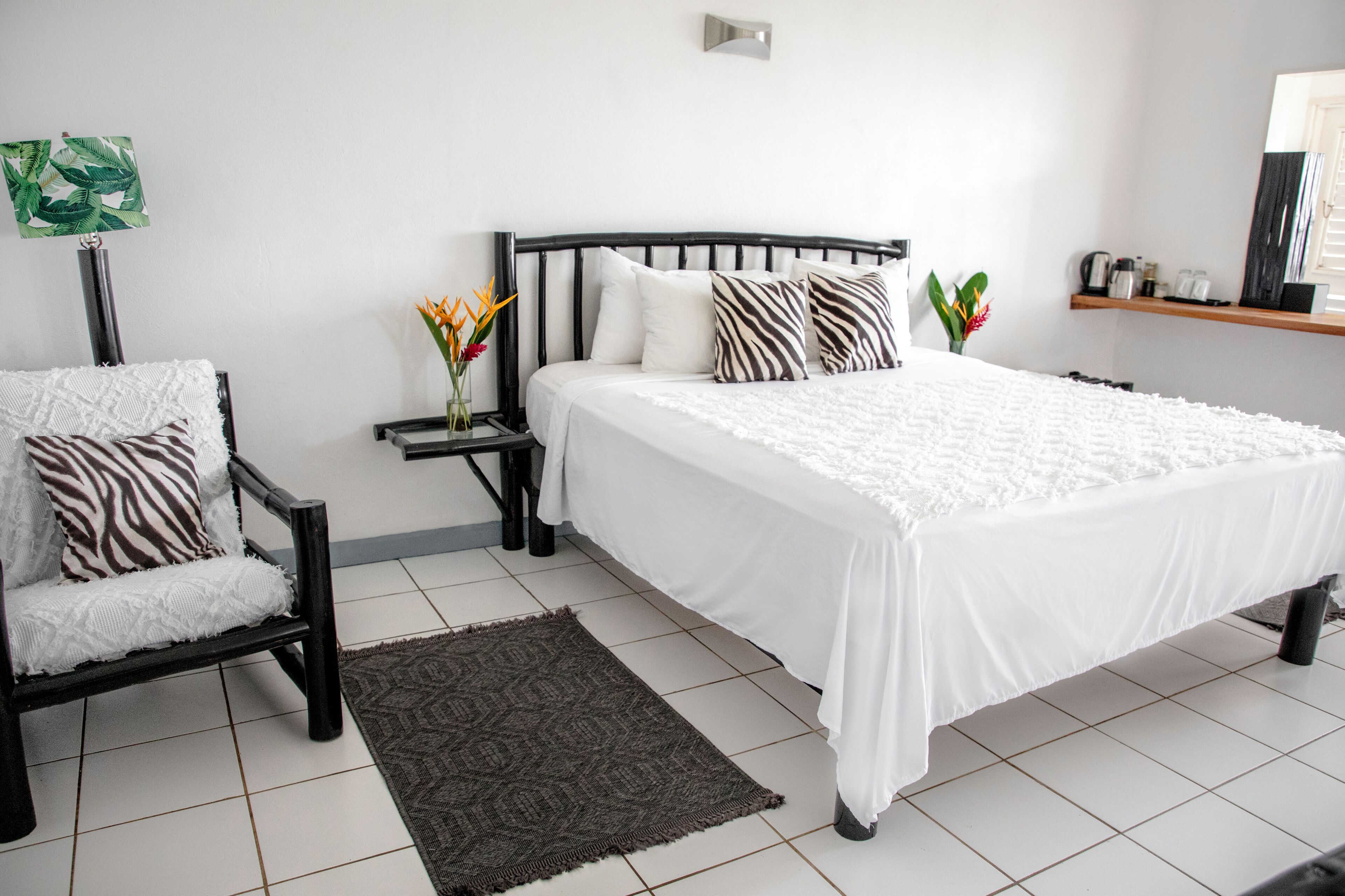 A bedroom with a large bed, a chair, a nightstand, a wall-mounted shelf, and a small table with a vase of flowers.