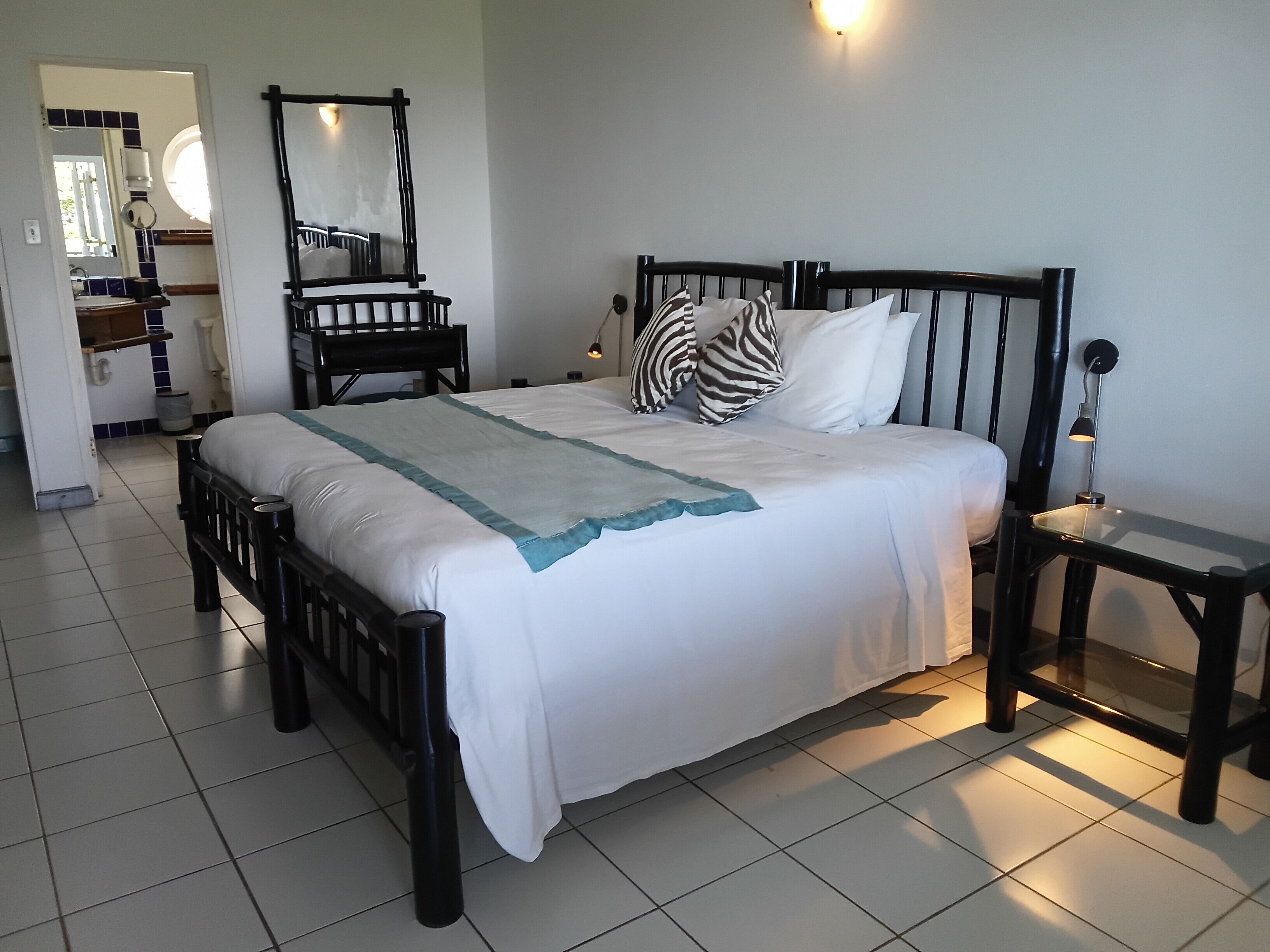 Un lit bien fait, recouvert d’une couette blanche et turquoise, agrémenté d’un coussin à motifs zébrés et doté d’une tête de lit en bois. On y trouve une petite table de chevet avec une lampe, ainsi qu’un grand miroir accroché au mur.