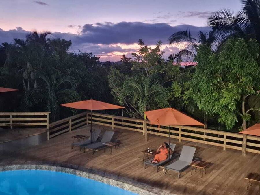 A poolside area with lounge chairs and umbrellas, surrounded by lush greenery and palm trees.