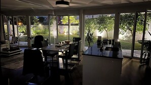 A modern kitchen with large windows, a dining area, and a view of a garden and street at night. - Garden apartment at Zurich lake (Richterswil)