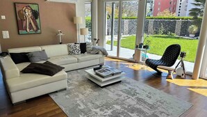 A modern living room with a white sofa, a coffee table with books, a black chair, and a large window offering a view of a garden and buildings. - Garden apartment at Zurich lake (Richterswil)