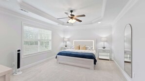 Une chambre à coucher comprenant un lit, un ventilateur de plafond, une grande fenêtre avec des stores, une table de chevet avec une lampe et un grand miroir.