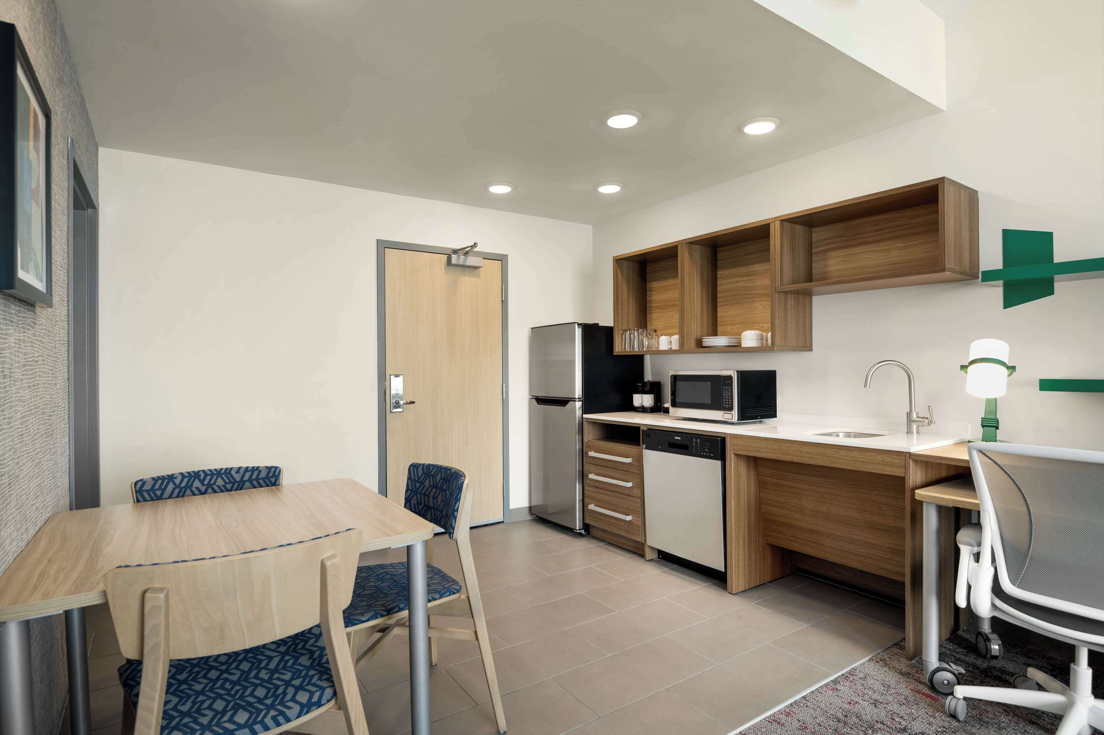 Una cocina compacta con encimera de madera, un refrigerador, un microondas y un lavamanos. Hay una mesa de comedor con sillas y una pequeña zona de trabajo con una silla y un escritorio.