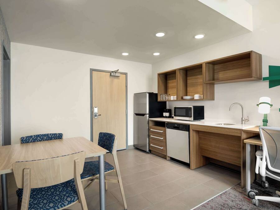 Una cocina compacta con encimera de madera, un refrigerador, un microondas y un lavamanos. Hay una mesa de comedor con sillas y una pequeña zona de trabajo con una silla y un escritorio.