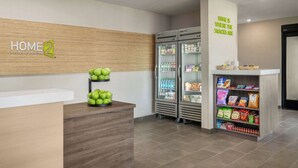 Una zona de recepción de hotel con un mostrador de madera, un cartel en la pared que dice "HOME 2 SUITES BY HILTON" y un estante de snacks con diferentes artículos.