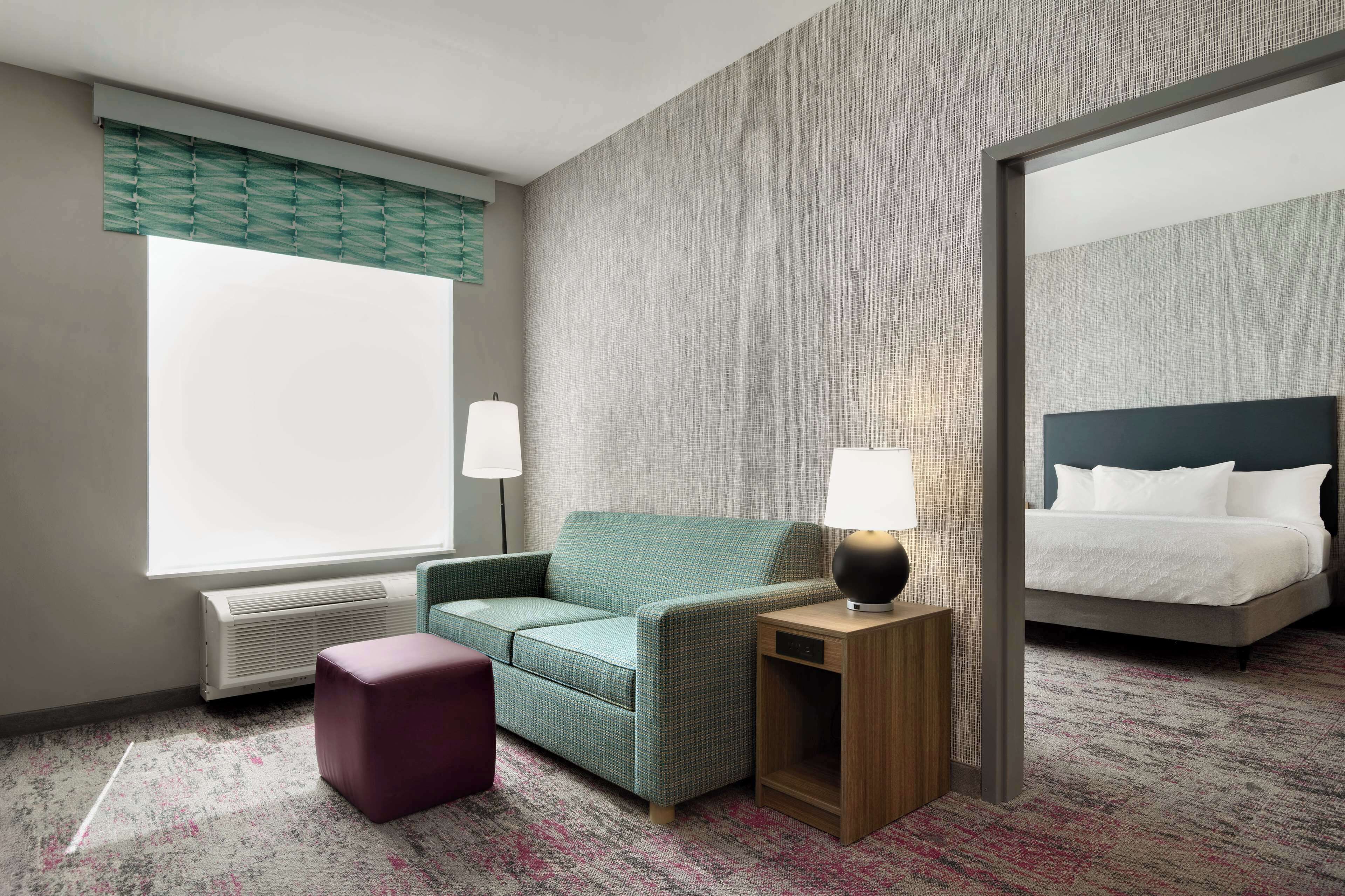 Habitación de hotel con sofá verde, una pequeña mesita de madera con lámpara, una cama grande con ropa blanca y un ventanal con persianas.
