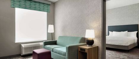 Habitación de hotel con sofá verde, una pequeña mesita de madera con lámpara, una cama grande con ropa blanca y un ventanal con persianas.