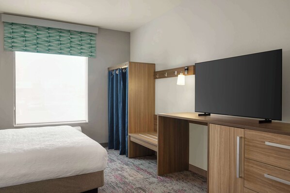A hotel room with a bed, a desk, a flat-screen TV, a window with curtains, and a wall-mounted light fixture.