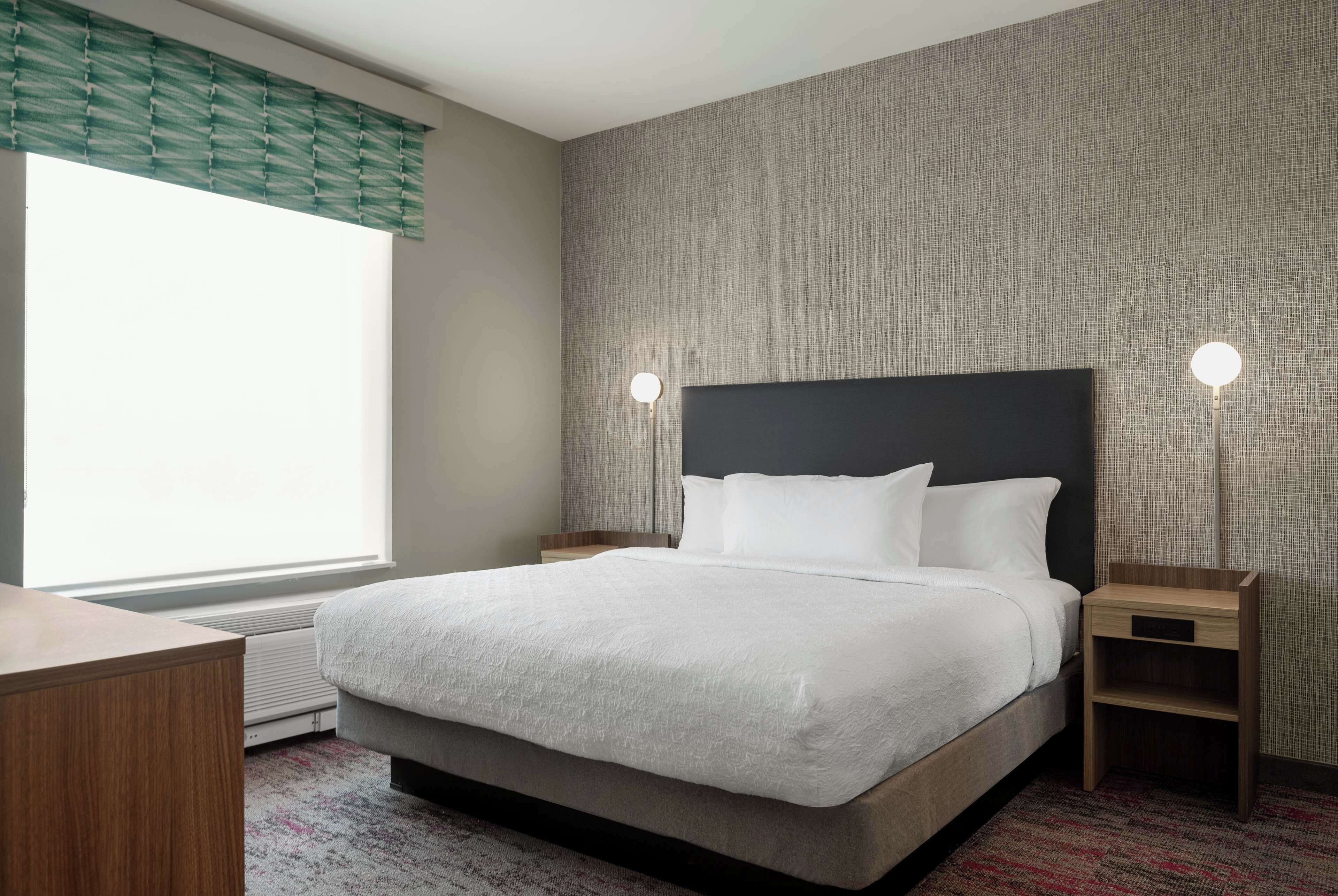 A modern hotel room with a large bed, a bedside table, a nightstand, a window with blinds, and a wall-mounted lamp.