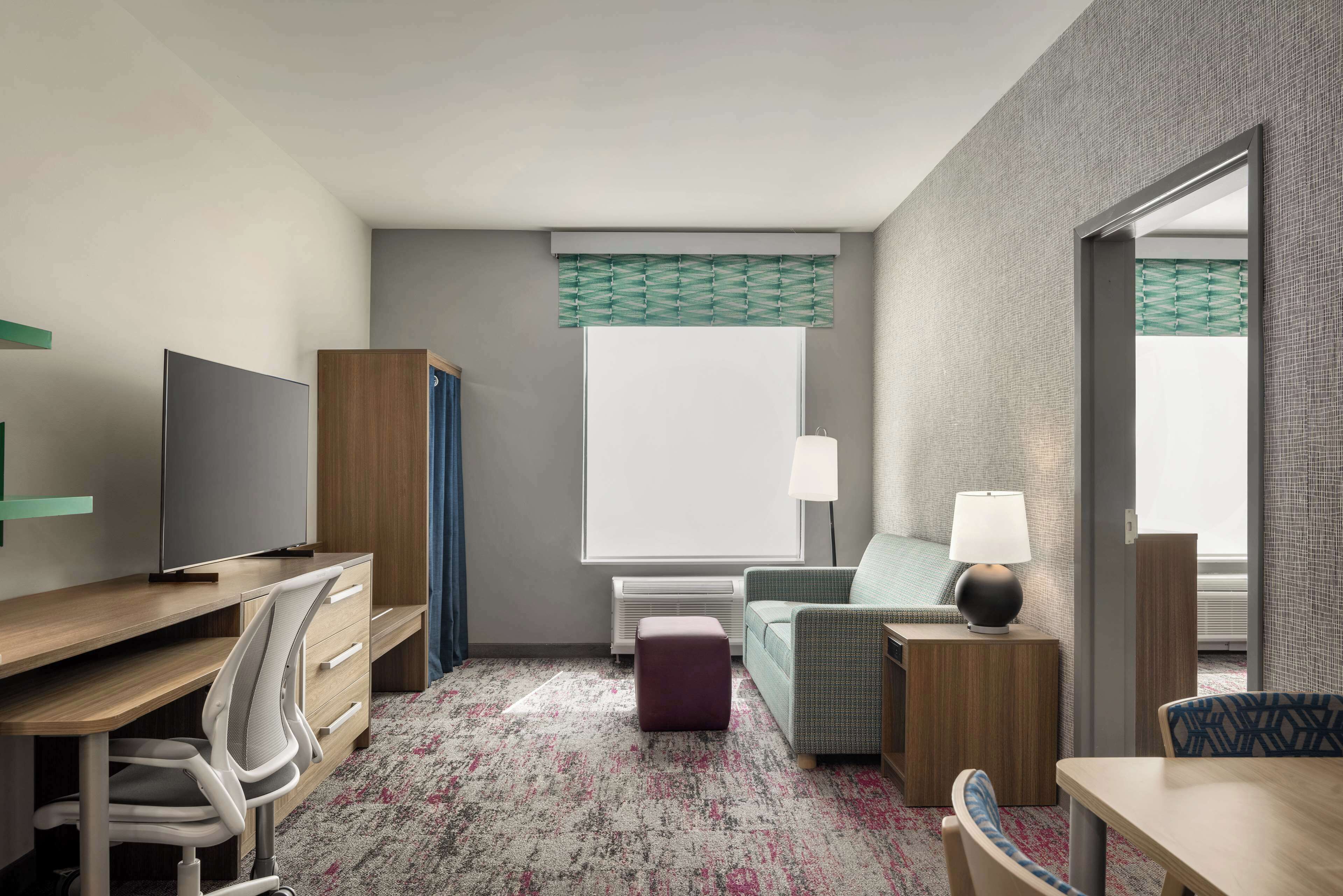 A hotel room with a desk, chair, bed, and a small table with a lamp.