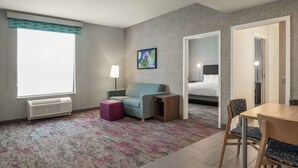 Una habitación de hotel con un ventanal grande, un sofá, una mesita, una lámpara, una cama y un comedor con sillas.