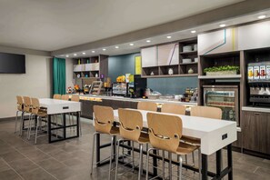 Un restaurante moderno con varias mesas y sillas, una barra con mostradores de comida y un refrigerador con bebidas.