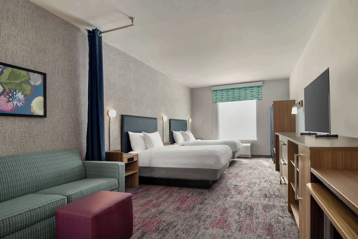 A hotel room with a sofa, two beds, a TV, a desk, and a window with curtains.