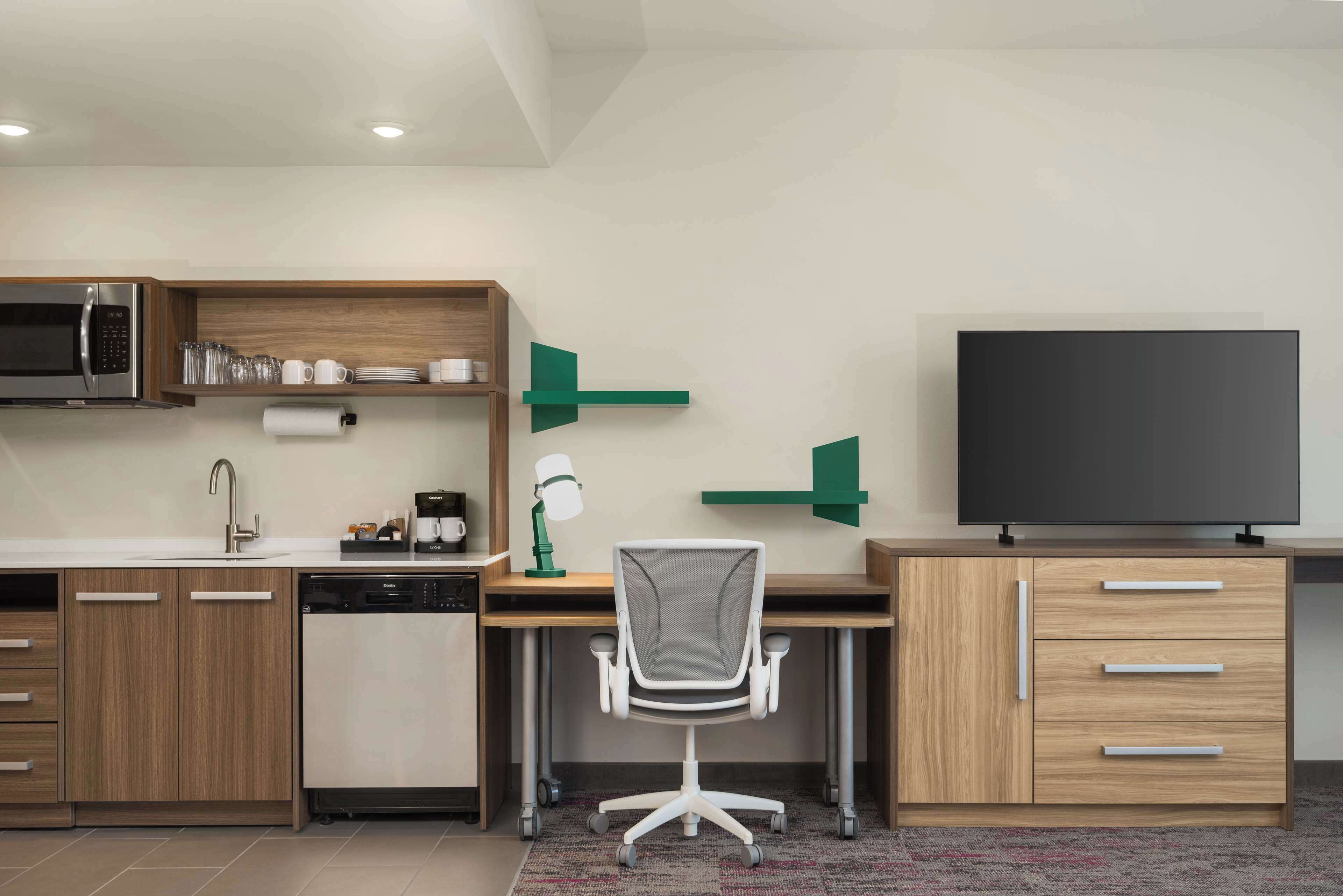 A modern kitchen with a built-in microwave, sink, and dishwasher. There is a wooden desk with a chair, a flat-screen TV, and a green decorative piece on the wall.