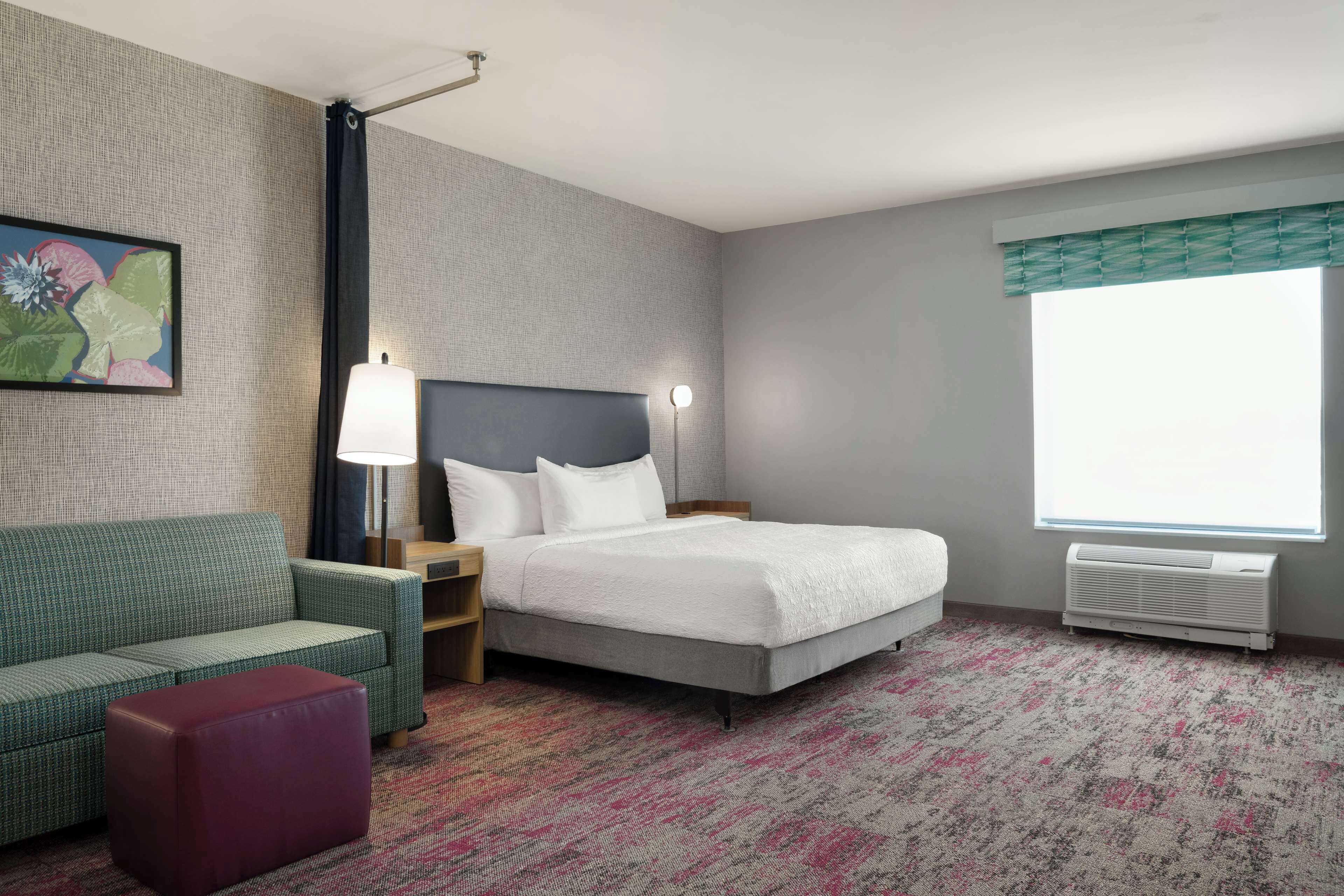 A hotel room with a bed, a sofa, a nightstand, a lamp, a window with blinds, and a radiator.