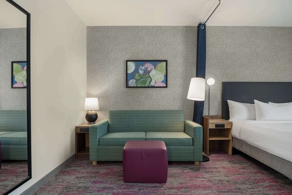 Habitación de hotel con sofá verde, un otoman morado, cama con sábanas blancas, mesita de noche con lámpara y una obra de arte enmarcada en la pared.