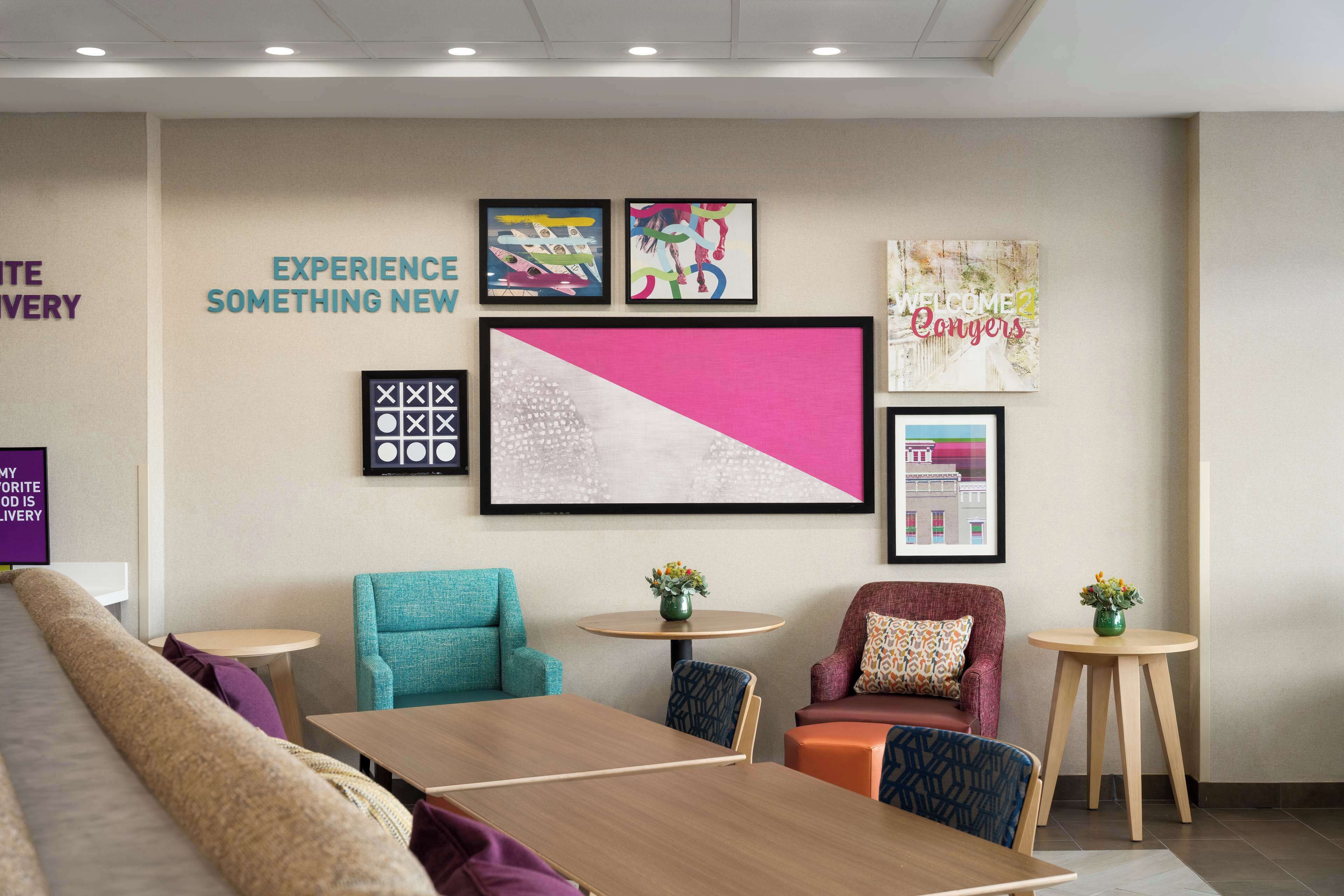 A modern hotel lobby with a long sofa, a wooden table, and two armchairs. There are framed artworks on the walls and a welcome sign.