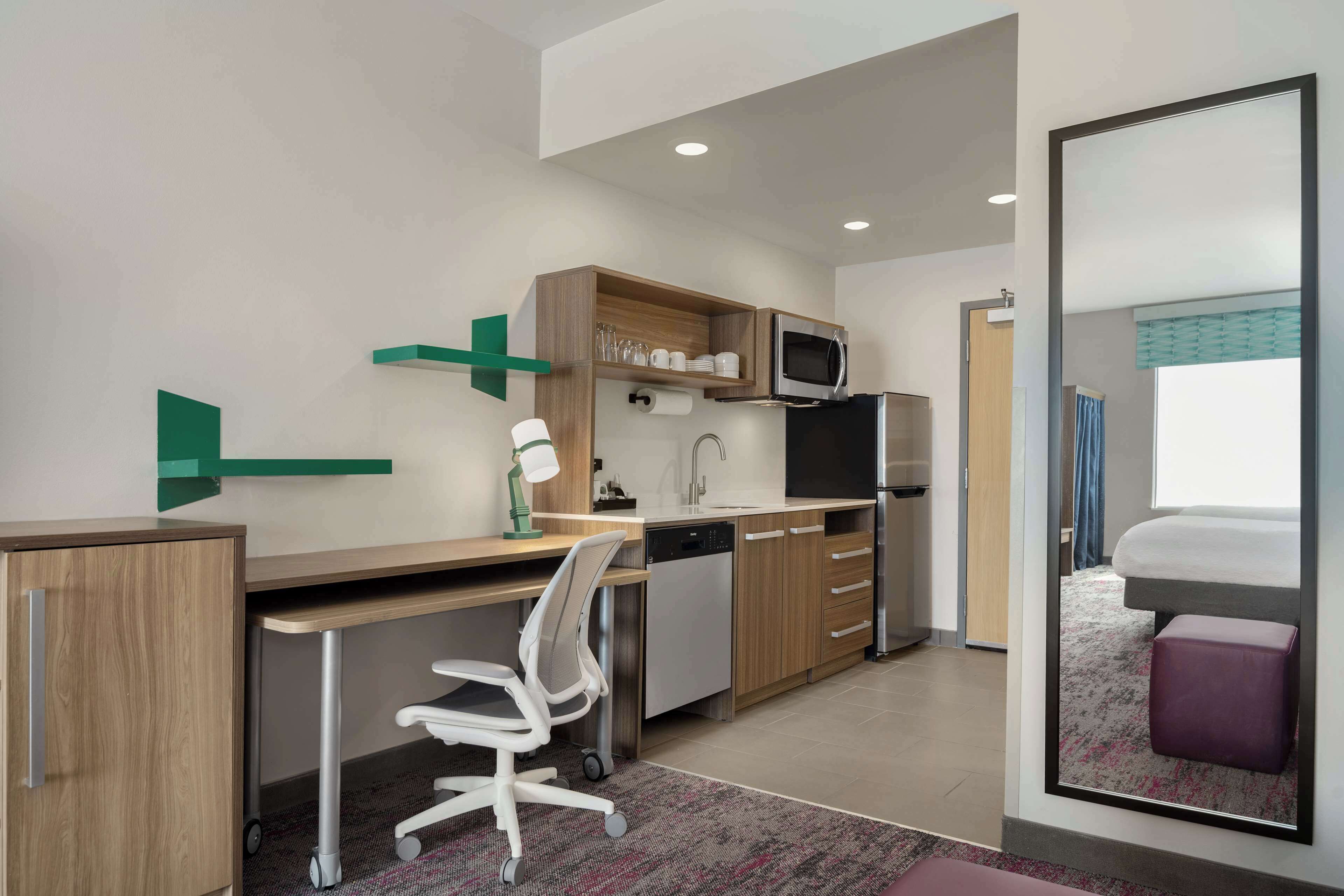 Quarto de hotel compacto com cozinha compacta, uma escrivaninha com cadeira, uma cama e um espelho grande.