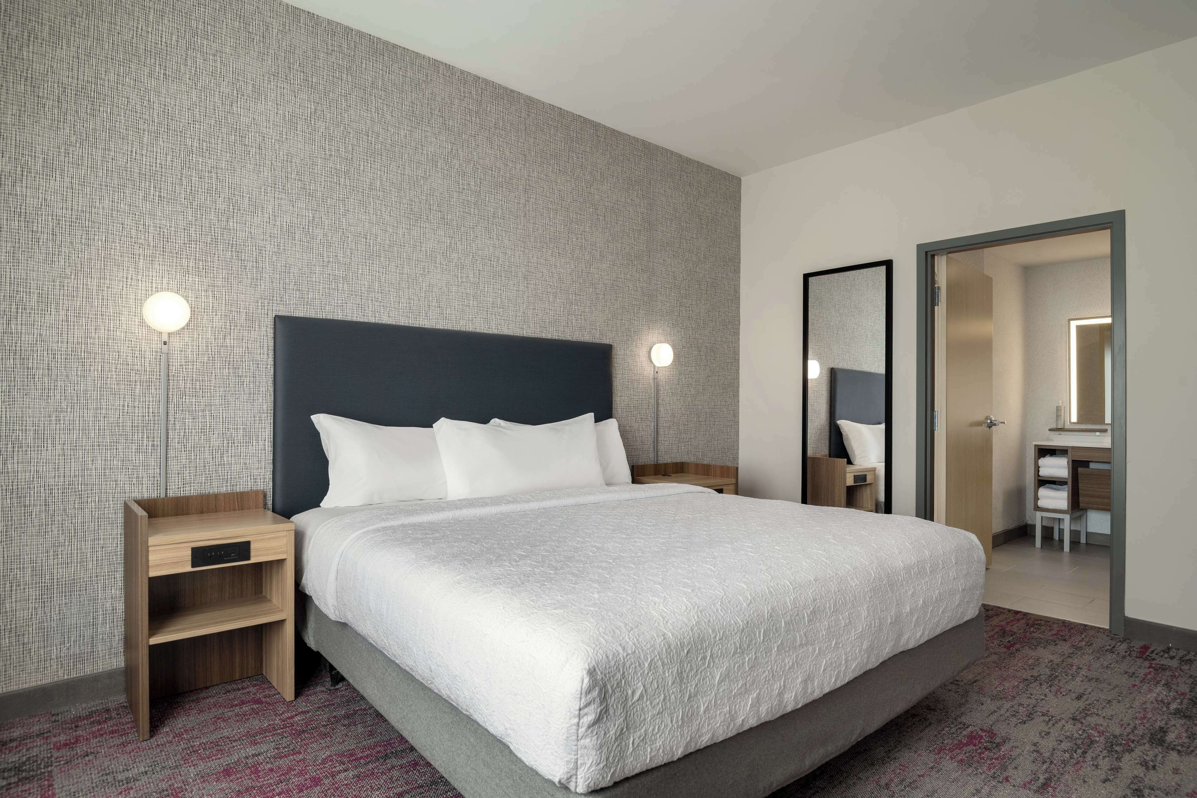Una habitación de hotel moderna con una cama grande, una mesita de noche de madera, un espejo y un baño visible a través de una puerta abierta.