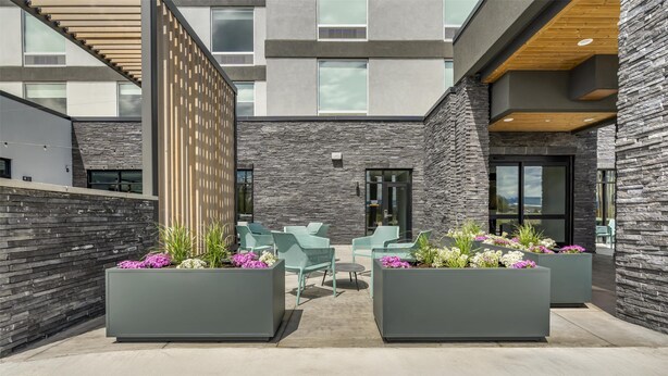 A modern outdoor seating area with planters and chairs, a stone wall, and a building entrance. - Home2 Suites By Hilton Bozeman Four Corners (Bozeman)