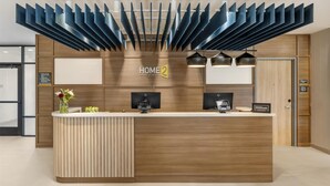 A modern reception desk with a wooden finish, a logo for 'HOME 2', and two black pendant lights above. - Home2 Suites By Hilton Bozeman Four Corners (Bozeman)