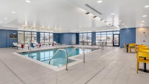A modern indoor swimming pool with clear glass walls, blue and white walls, and yellow chairs. - Home2 Suites By Hilton Bozeman Four Corners (Bozeman)