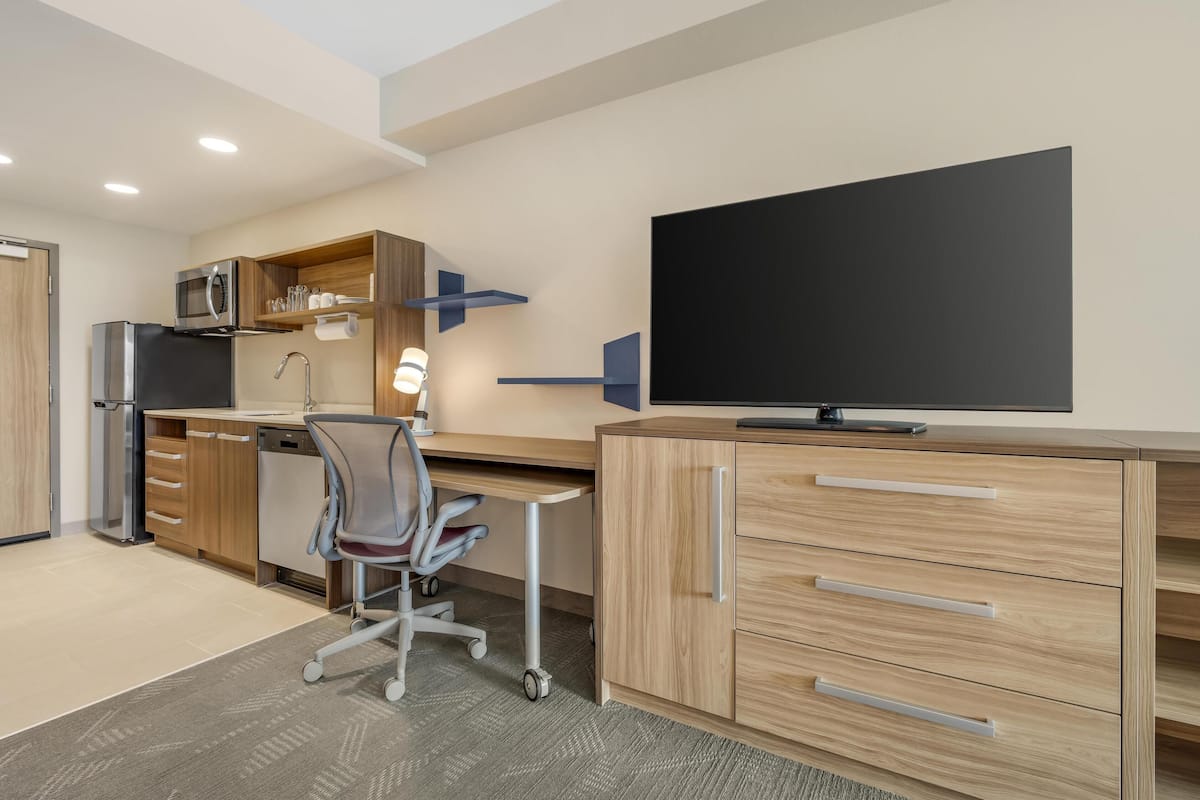 A modern kitchen with a microwave, refrigerator, and sink. There is a wooden desk with a chair and a television on top.