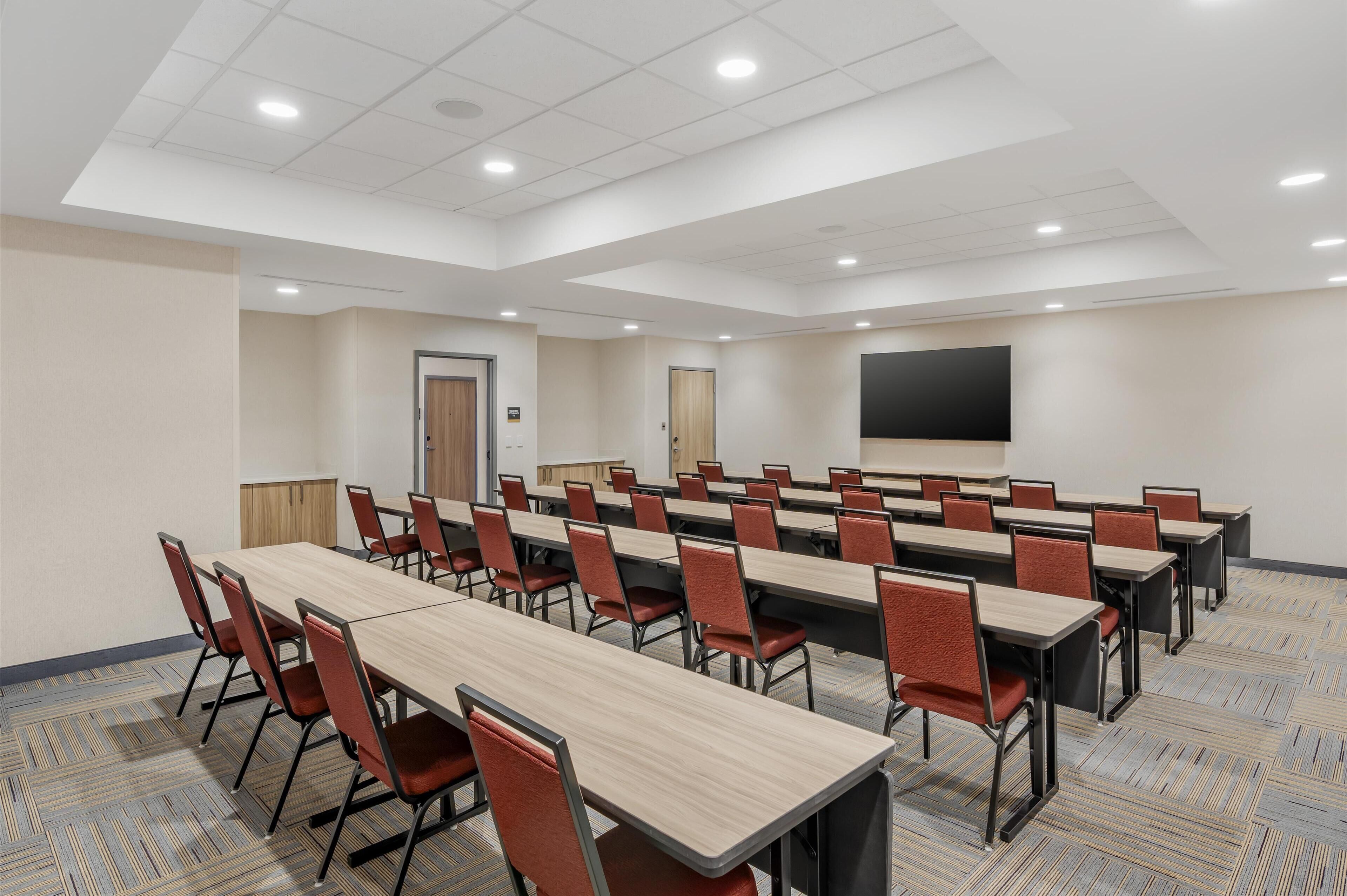 Une salle de conférence dotée de plusieurs tables et chaises, d’un grand écran et d’un sol recouvert de moquette.
