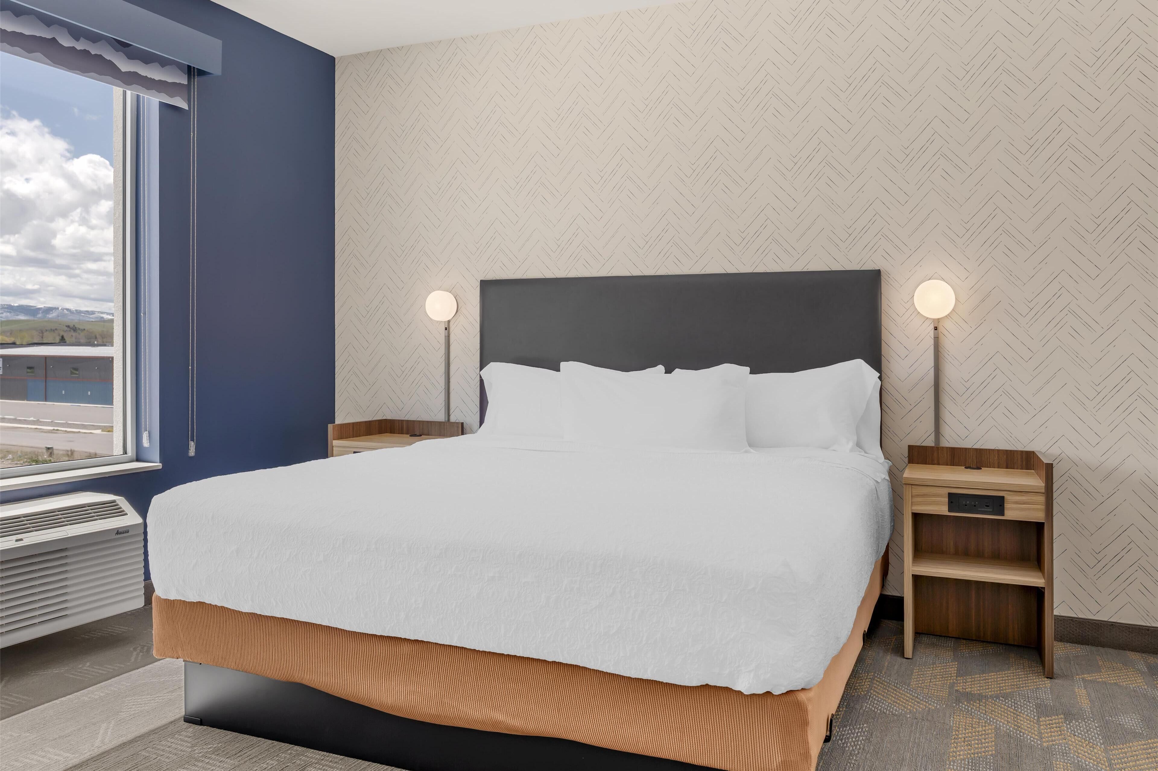 A neatly made bed with white linens, two bedside tables with lamps, a window with a view, and a patterned wall.