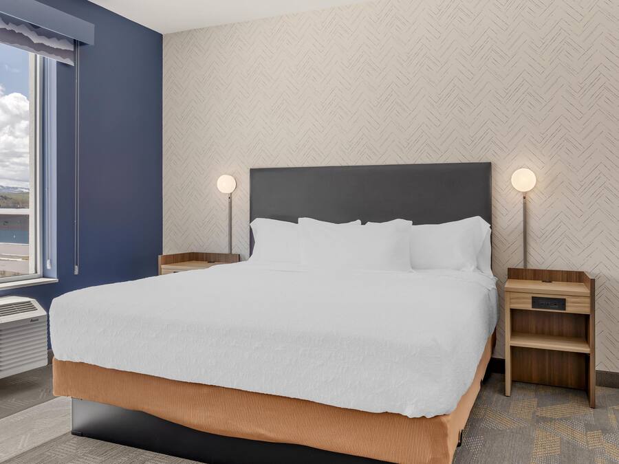 A neatly made bed with white linens, two bedside tables with lamps, a window with a view, and a patterned wall.