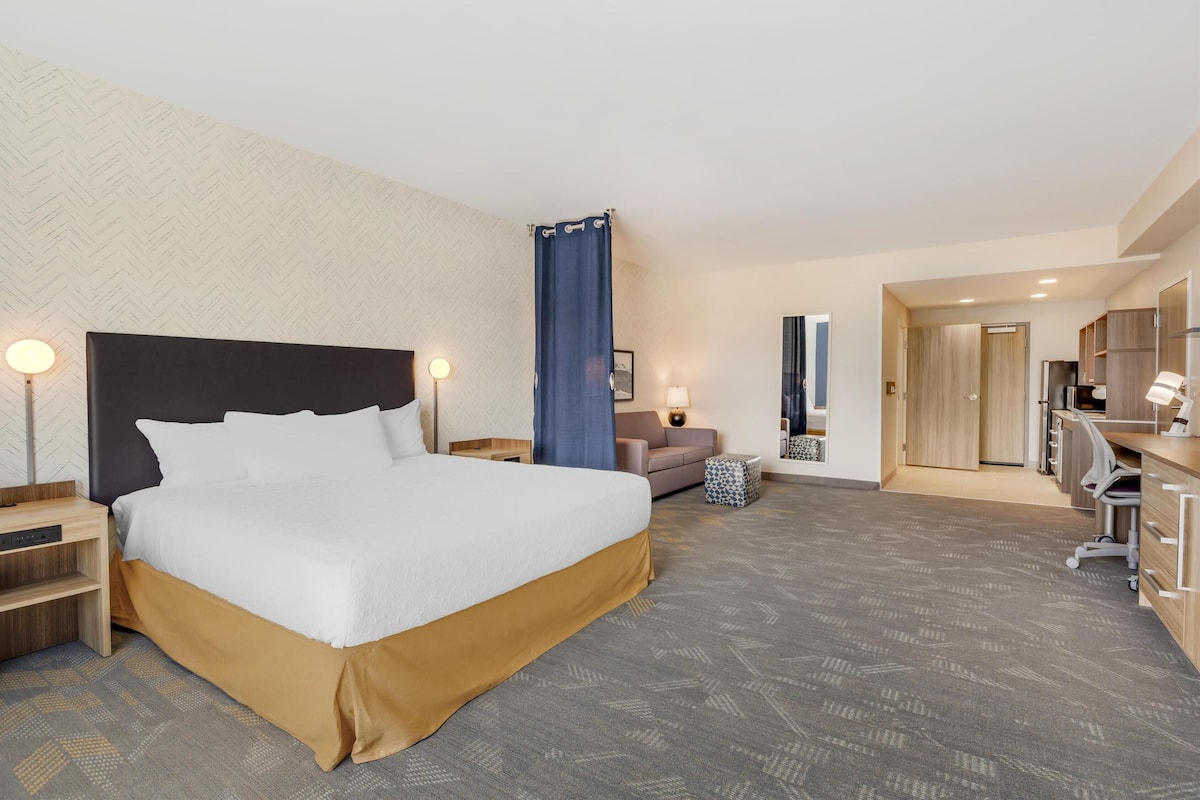 A hotel room with a large bed, a desk, a chair, a sofa, and a bathroom visible through an open door.