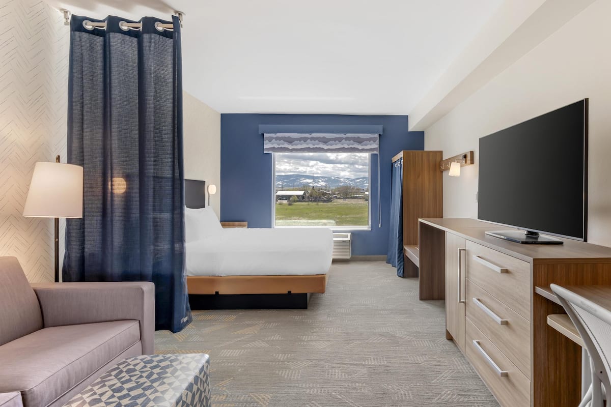 A hotel room with a bed, a TV, a desk, and a view of a mountainous landscape.