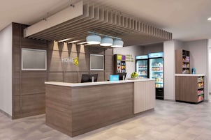 A modern hotel reception area with a wooden reception desk, a digital display, and a minibar.