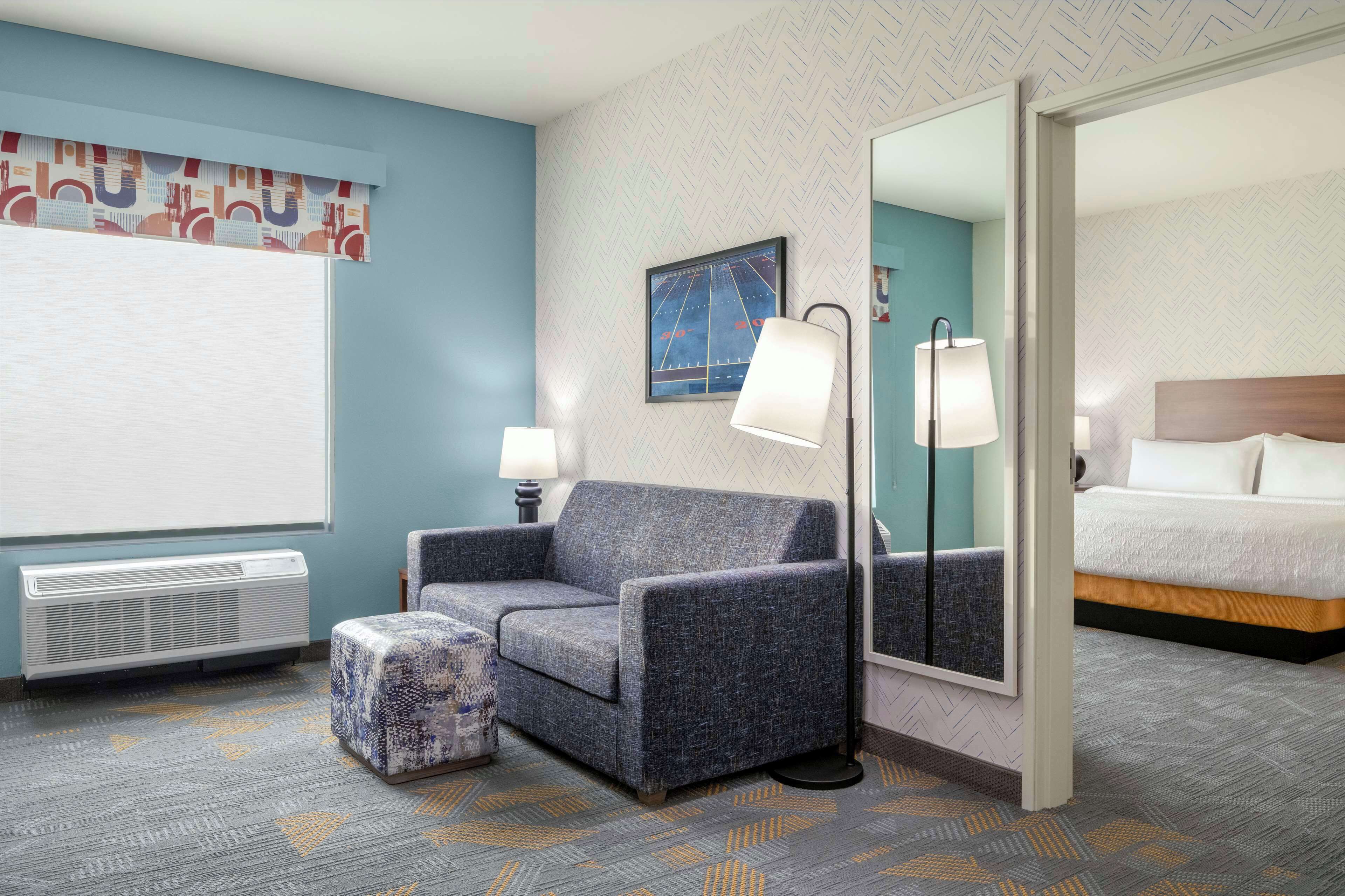 A hotel room with a blue wall, a large window with a white blind, a grey sofa, a matching ottoman, a floor lamp, a bed with white bedding, and a mirror reflecting the room.