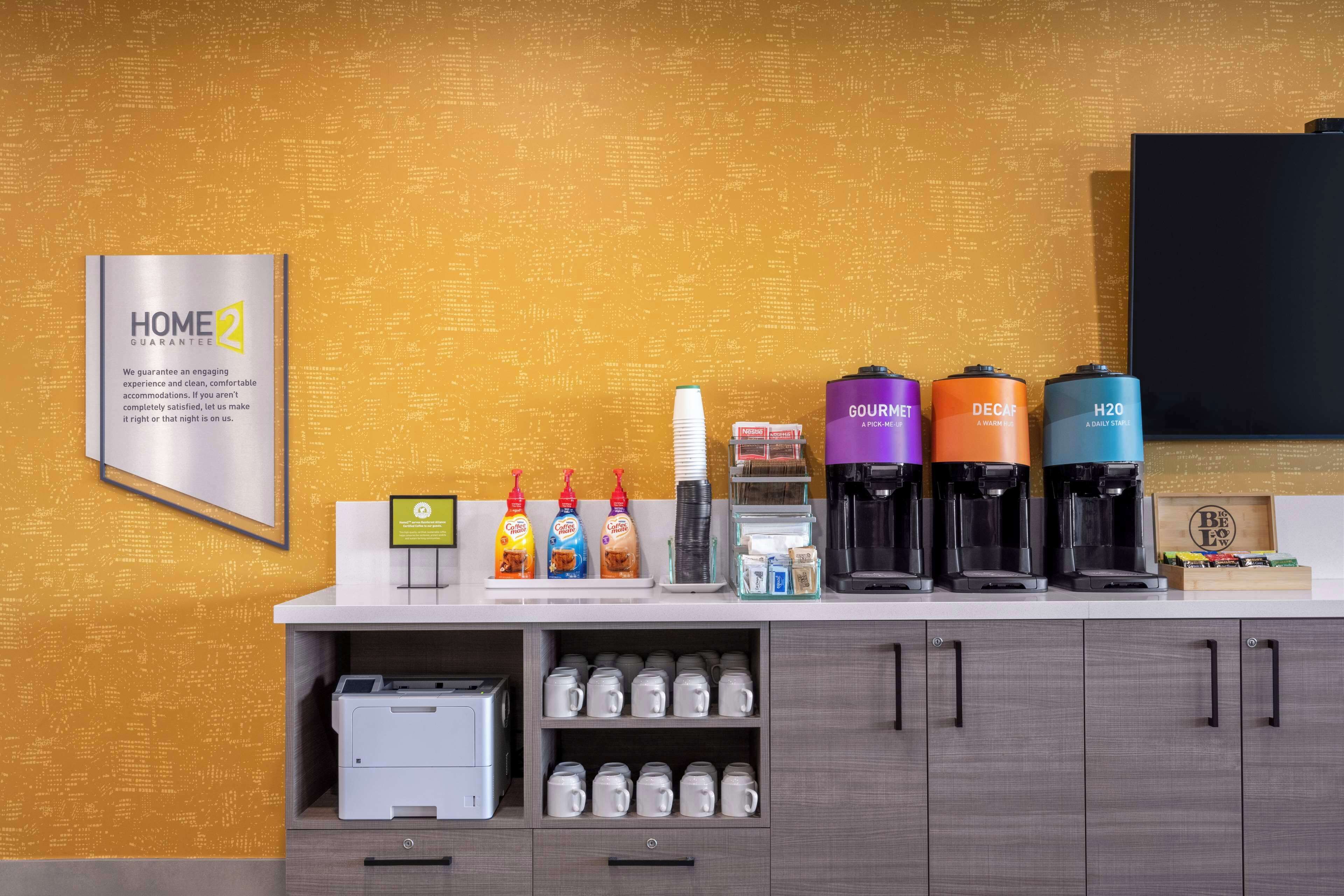 Een hotel receptie met een 'HOME2 GUARANTEE' bord, koffiezetapparaten, drankdispensers en een printer.