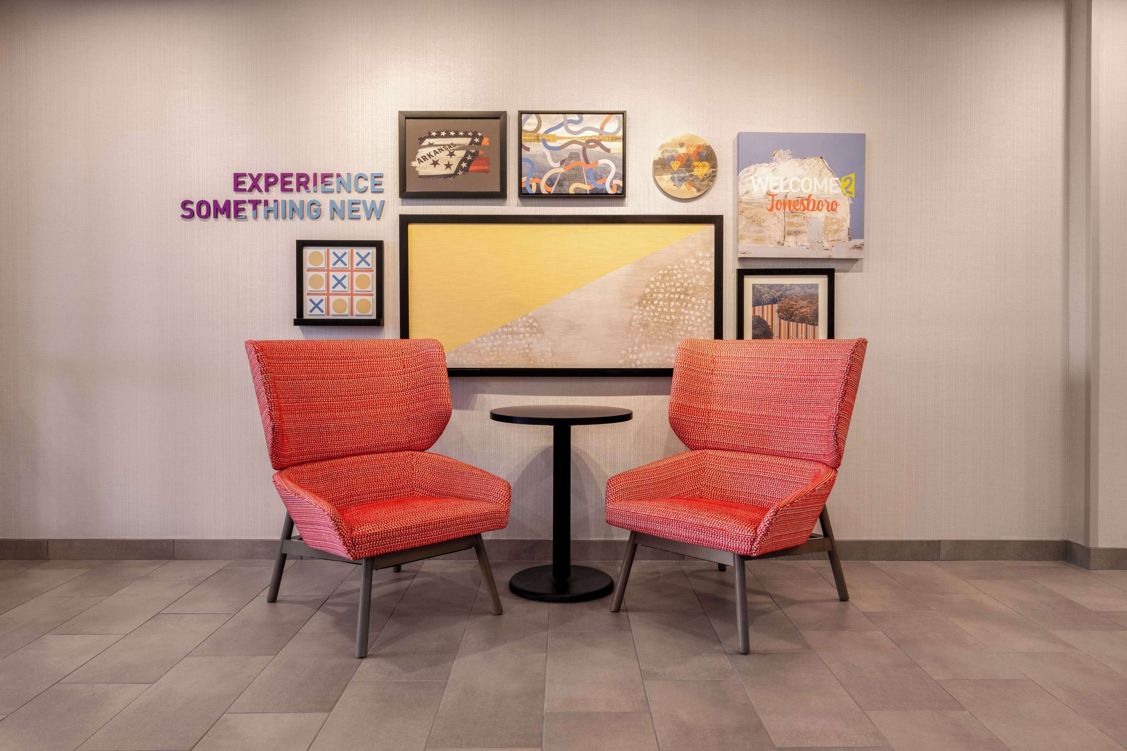 A room with two red chairs and a small round table, a wall with framed artwork, and a sign that reads "WELCOME Jonesboro".