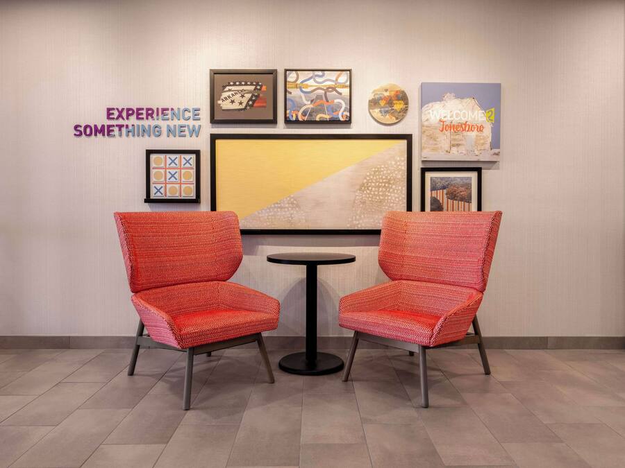 A room with two red chairs and a small round table, a wall with framed artwork, and a sign that reads "WELCOME Jonesboro".