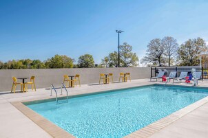 Una piscina rectangular con borde de azulejos, rodeada por una terraza de color claro. Hay sillas y mesas amarillas dispuestas a lo largo de la piscina, y una zona de estar cubierta con sillas azules y blancas.