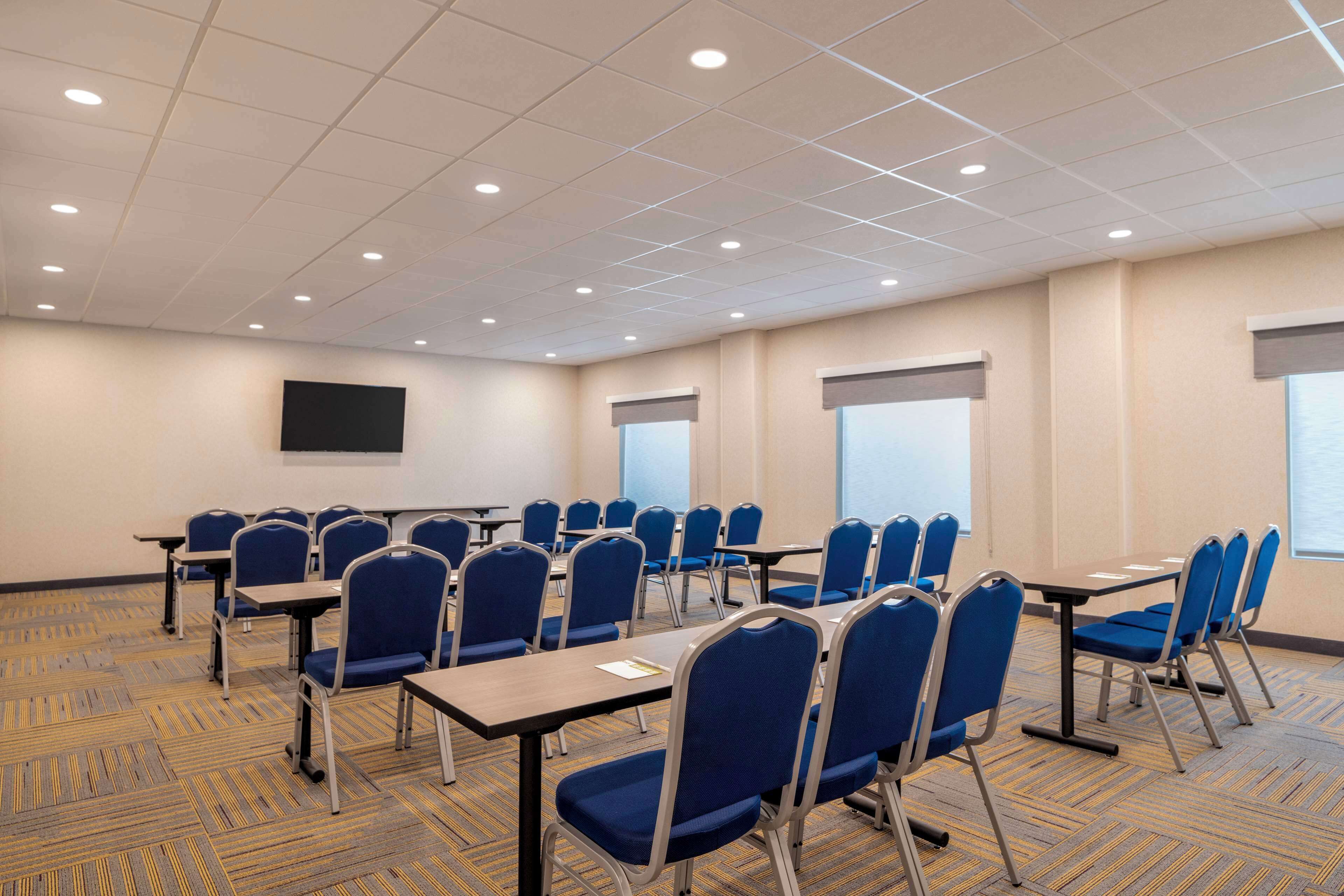 Un salón de conferencias con filas de sillas azules y mesas, un televisor de pantalla plana en la pared y ventanas amplias con persianas.