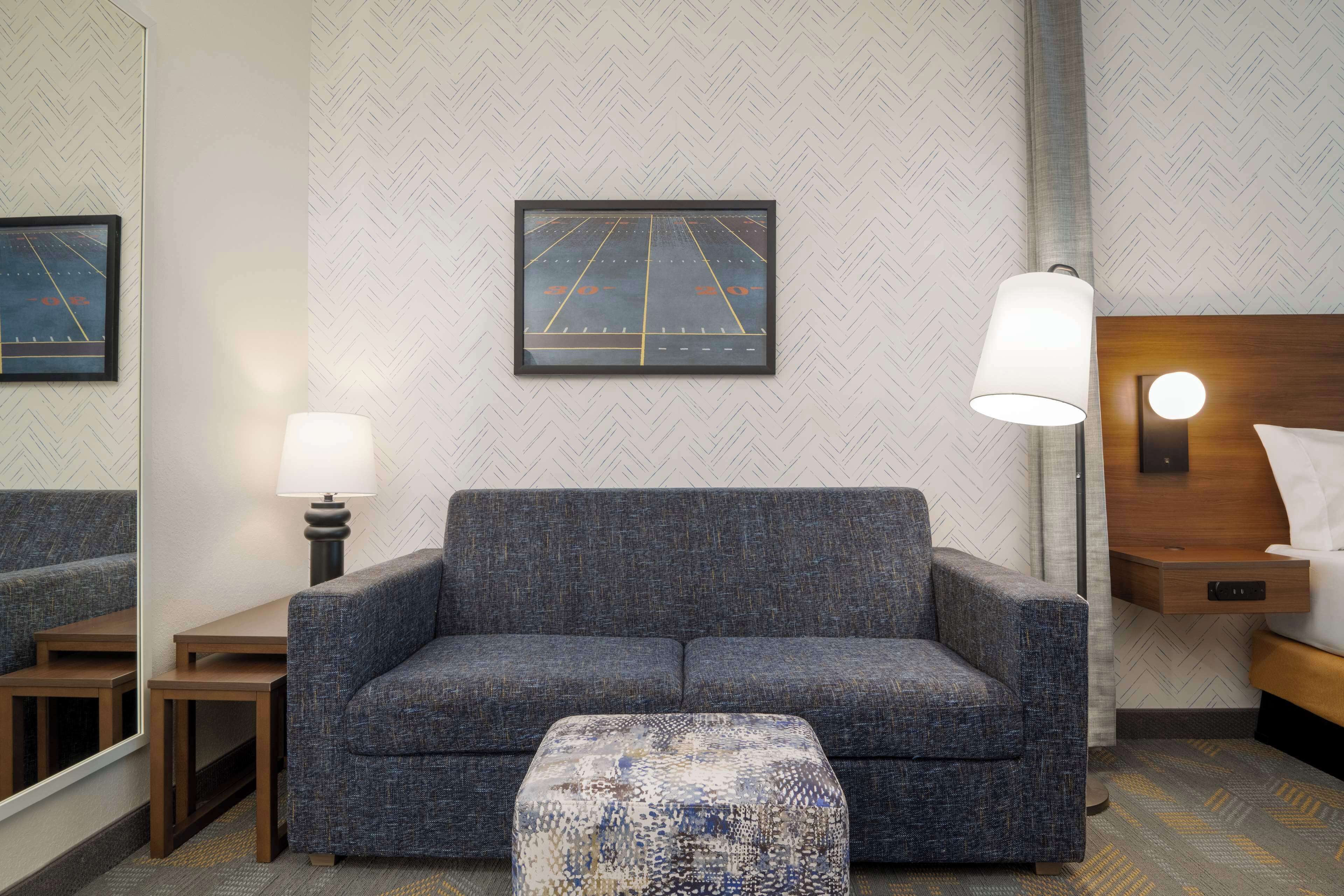 A hotel room with a gray sofa, a small table, a lamp, a bed with a wooden headboard, and a mirror on the wall.