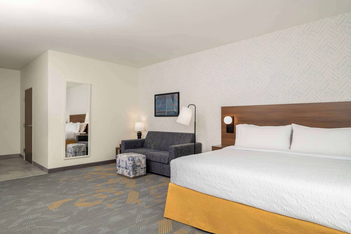 Habitación de hotel con una cama grande, un sofá, un pequeño reposapiés, un espejo y un cuadro en la pared.