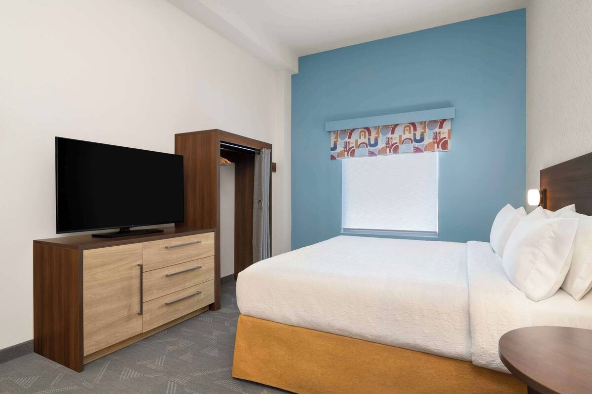 Habitación de hotel con una cama, un televisor sobre un tocador de madera, una ventana con cortinas y una pared de acento azul.