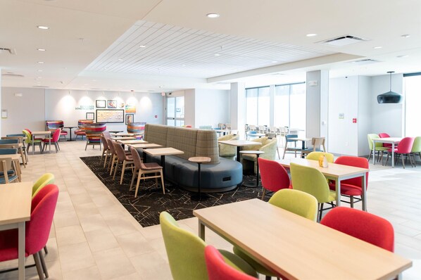 A modern dining area with multiple tables and colorful chairs, featuring a central seating area with a cushioned bench and a black and white patterned rug. - Home2 Suites By Hilton Effingham (Effingham)