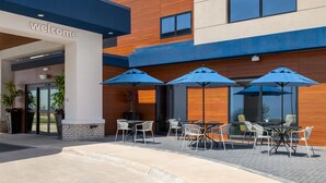 A modern building with a 'welcome' sign, outdoor seating with umbrellas, and a brick pathway.