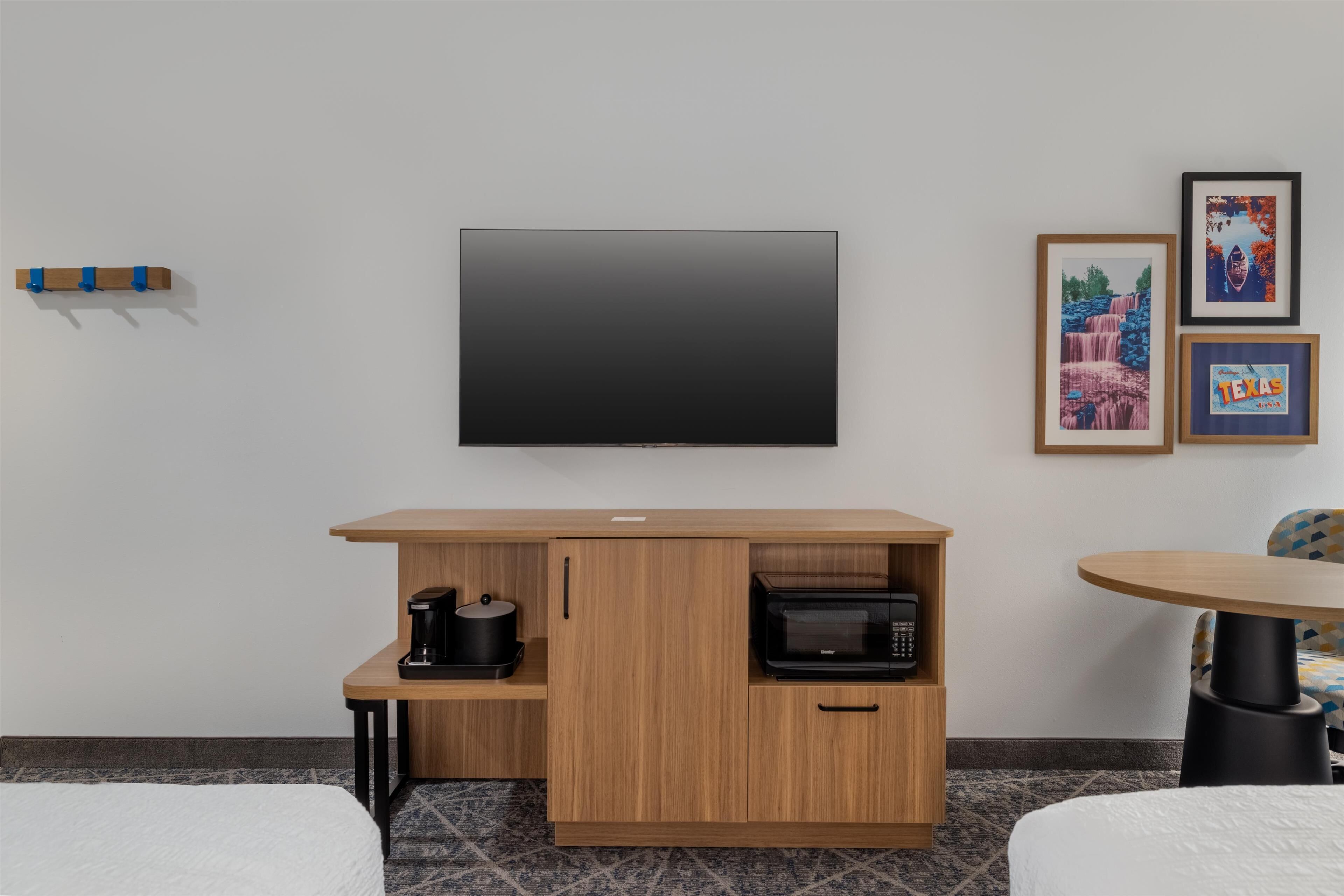 Habitación de hotel con televisión de pantalla plana montada en la pared, un mueble de madera con cafetera y microondas, una mesita con una silla y cuadros enmarcados en la pared.