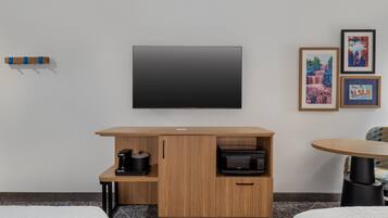 Habitación de hotel con televisión de pantalla plana montada en la pared, un mueble de madera con cafetera y microondas, una mesita con una silla y cuadros enmarcados en la pared.